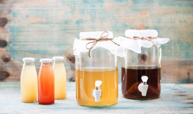 Kombucha em três momentos: nas garrafinhas, após a segunda fermentação. Na jarra com líquido mais claro, a bebida pronta após primeira fermentação. Ao fundo, o chá com o scoby ou "panqueca" no primeiro dia de fermentação. Foto: Letícia Akemi/Gazeta do Povo