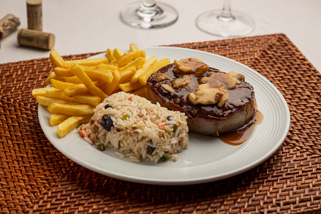 Medalhão Fornão, um dos clássicos do tradicional O Fornão, traz filé mignon grelhado com bacon, coberto com molho madeira e champignon, servido com arroz à grega e batatas fritas.