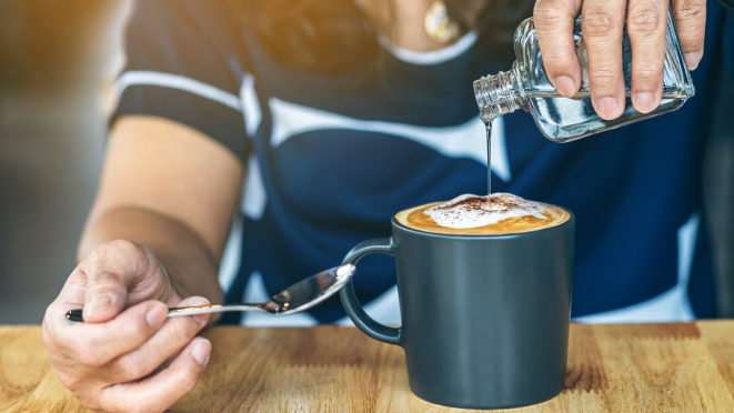 Close up of mature woman pouring warm syrup into hot cappuccino on cafe table. Pouring syrup in to hot coffee cup. Adding a subtle sweetness to favorite cup of coffee is delicious. Selective focus.