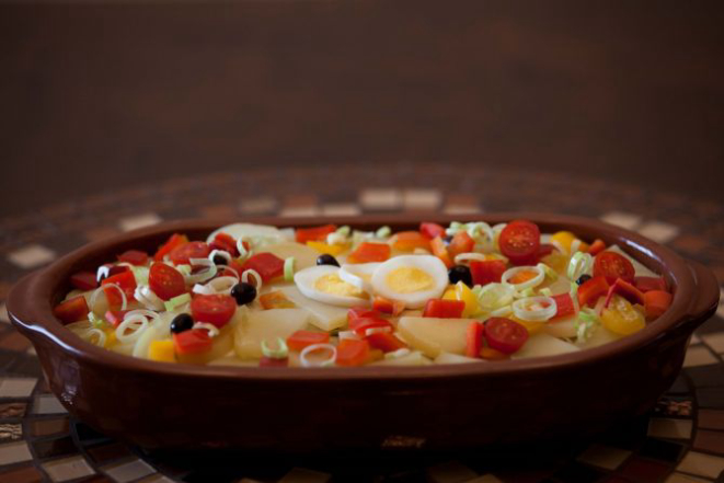 Lombos de bacalhau disposto em camadas com cebolas refogadas, pimentões, tomates cereja, alho poró, azeitonas pretas, ovos cozidos e batatas fatiadas. Foto: Divulgação