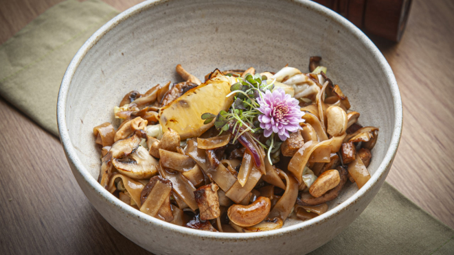 Serve a versão vegana do pad thai veggi composto por talharim de arroz, tofu defumado, mix de legumes, cogumelos e castanhas de caju.