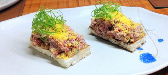 O Tartar de carne vem sobre uma camada de brioche na brasa, com creme de cebola e gema curada ralada. | Foto: Anacreon de Téos